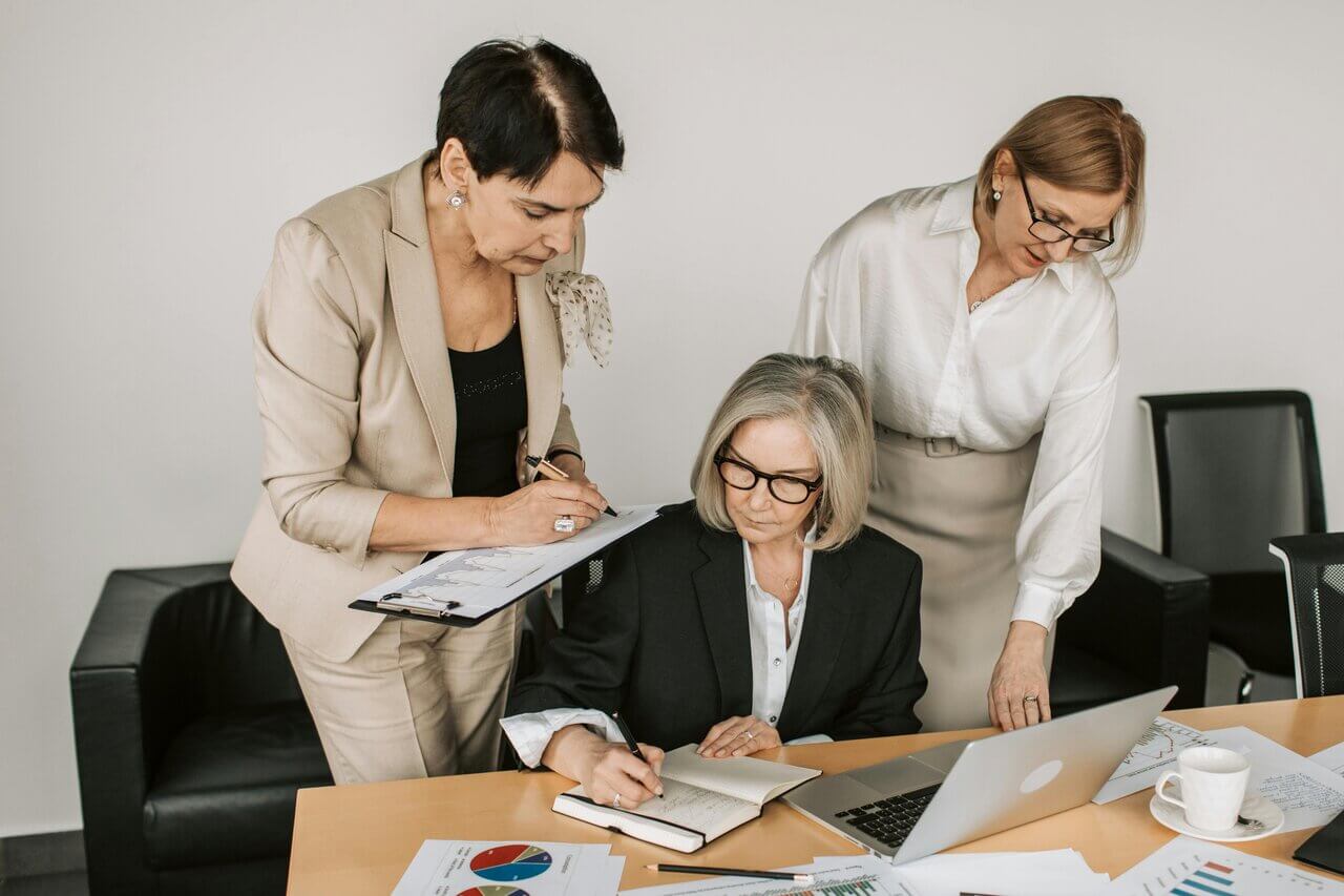 Three female professionals at desk in office with notebook and laptop